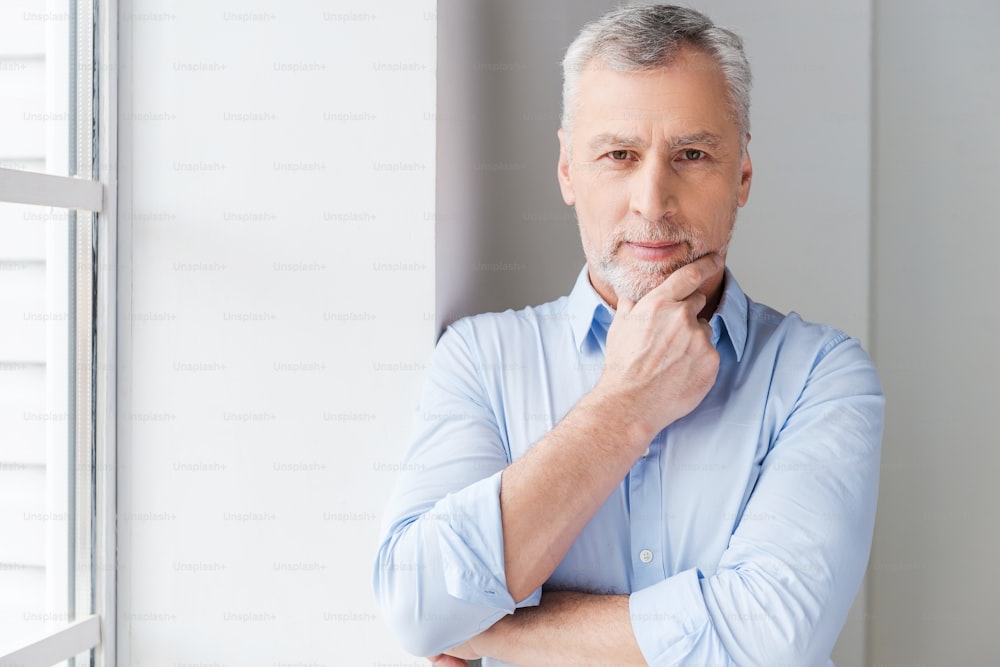 Thoughtful grey hair senior man in shirt looking away while standing near the window