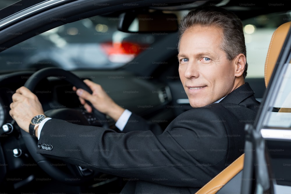 Side view of cheerful senior man in formalwear sitting in car and looking over shoulder