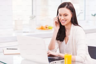 Attractive young woman working on laptop and talking on the mobile phone while sitting in the kitchen