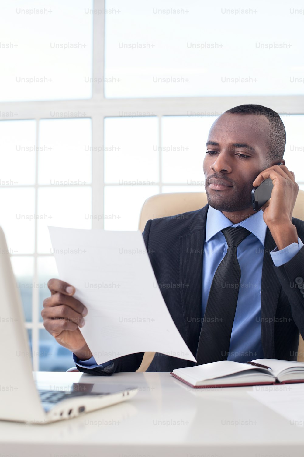 Selbstbewusster junger afrikanischer Geschäftsmann telefoniert und prüft Dokumente, während er an seinem Arbeitsplatz sitzt