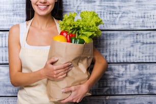 Cropped image of beautiful young woman in apron holding paper shopping bag full of fresh vegetables and smiling while standing in front of wooden background