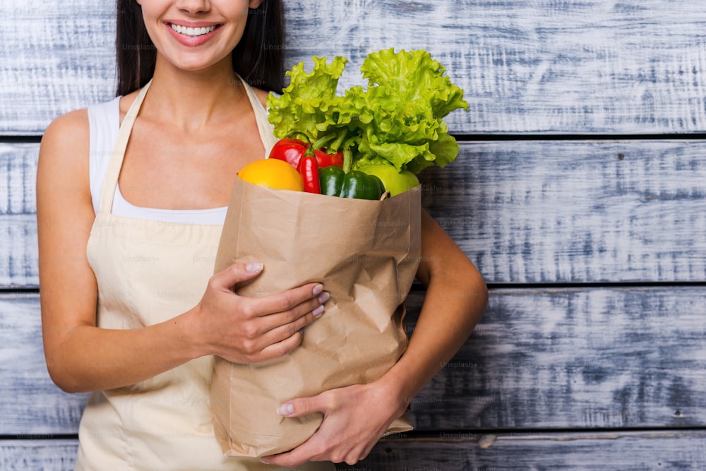 Imagem recortada da bela mulher jovem no avental segurando saco de compras de papel cheio de legumes frescos e sorrindo enquanto está em pé na frente do fundo de madeira