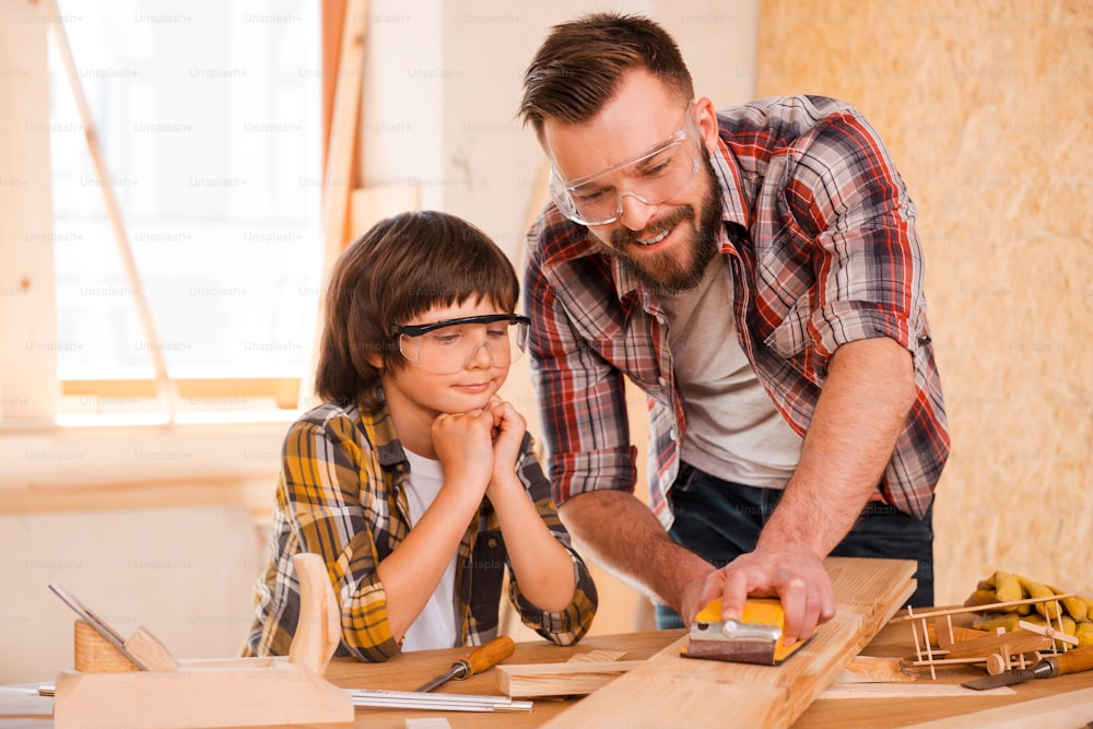 Smiling young male carpenter showing his son how to sand wood in his workshop