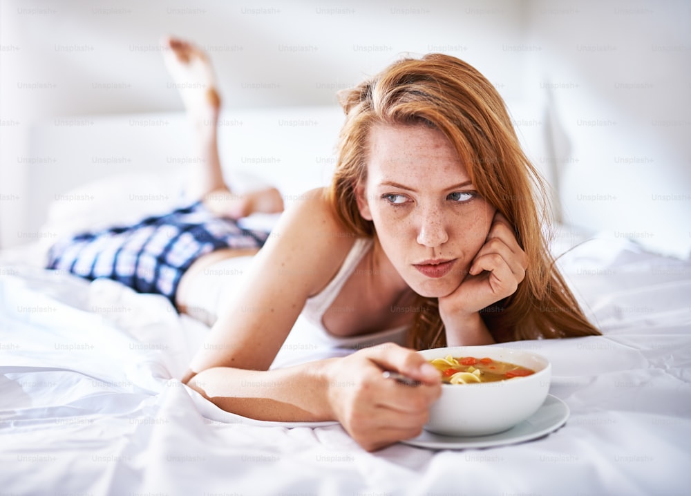 Frau im Bett isst Hühnernudelsuppe, während sie krank zur Seite schaut