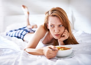 woman in bed eating chicken noodle soup while sick looking off to the side
