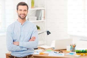 Handsome young man in shirt keeping arms crossed and smiling at camera while leaning at the desk in office