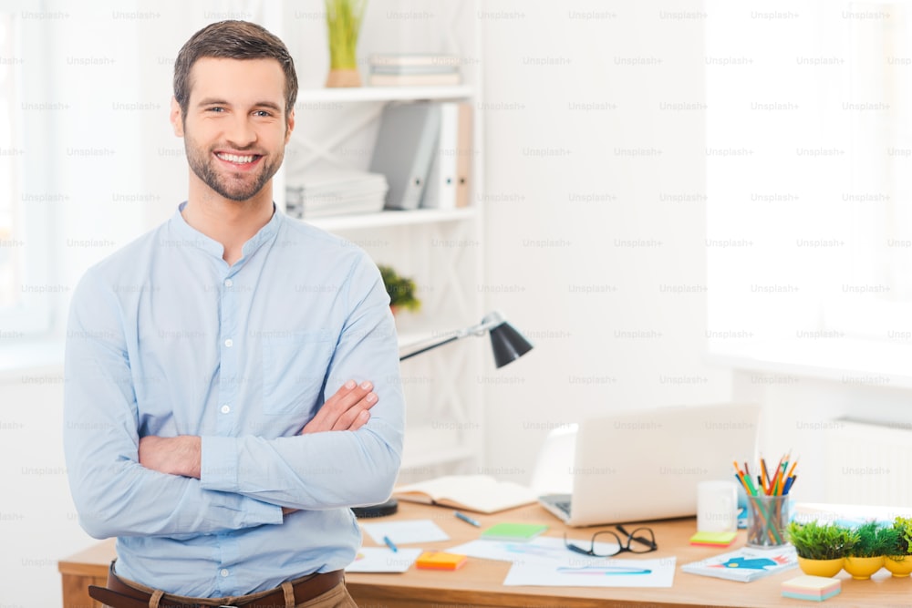 Beau jeune homme en chemise gardant les bras croisés et souriant à la caméra tout en se penchant au bureau dans le bureau