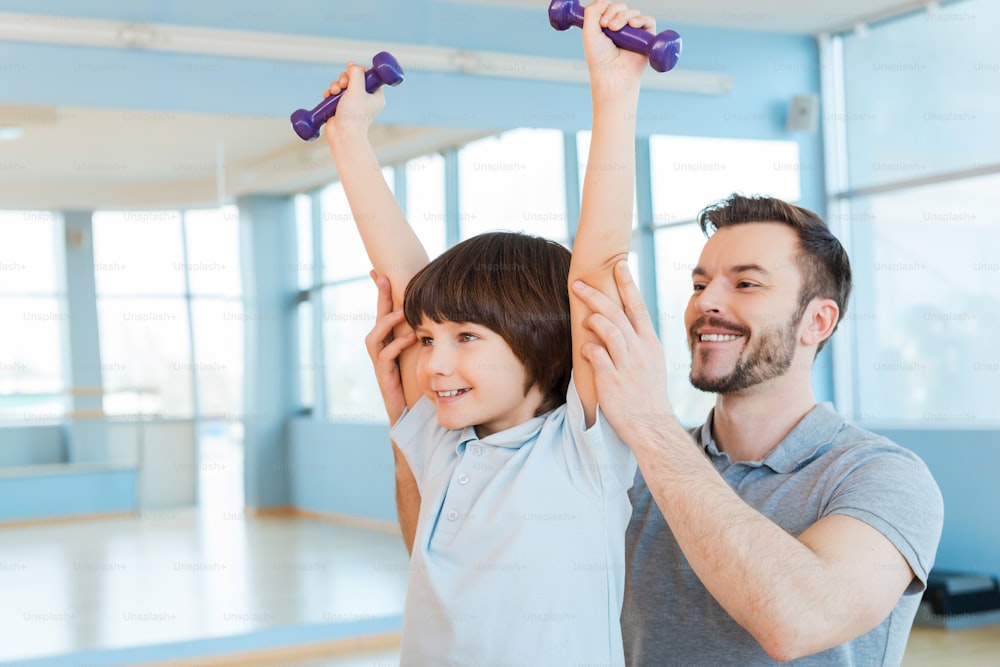 Padre feliz apoyando a su hijo en el entrenamiento con pesas mientras ambos están de pie en el gimnasio