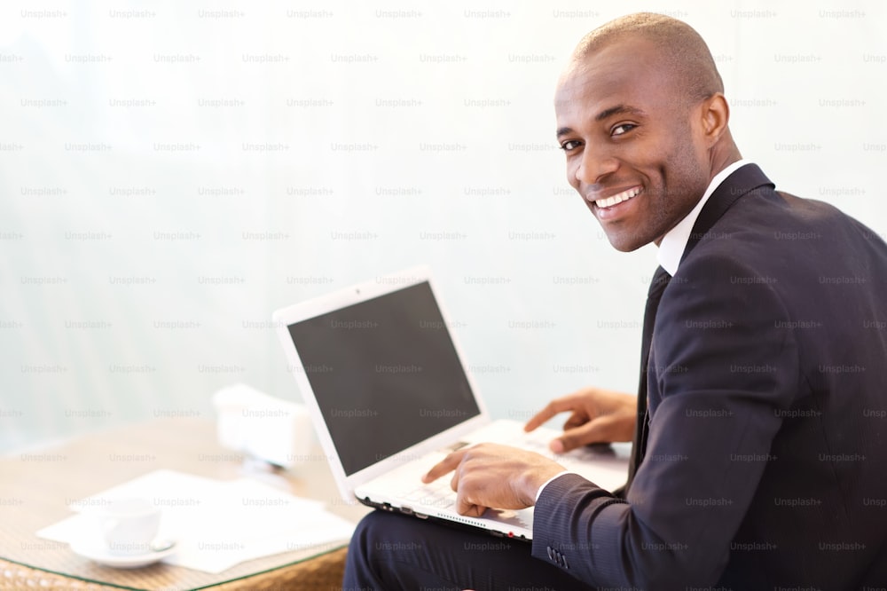Jovem empresário africano alegre digitando algo no laptop e olhando por cima do ombro