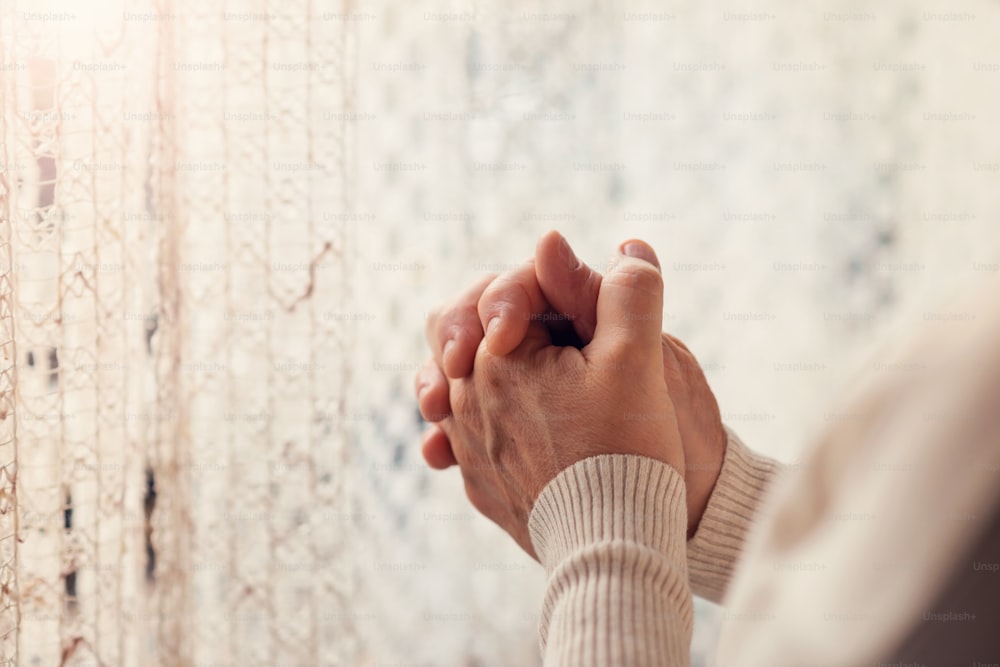 Hands of an unrecognizable woman standing by the window and praying