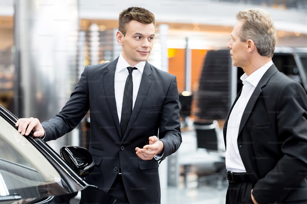 Handsome young classic car salesman standing in the dealership and helping a client to make a decision about car