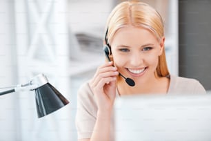 Mujer joven feliz que trabaja en la computadora y ajusta sus auriculares mientras está sentada en su lugar de trabajo