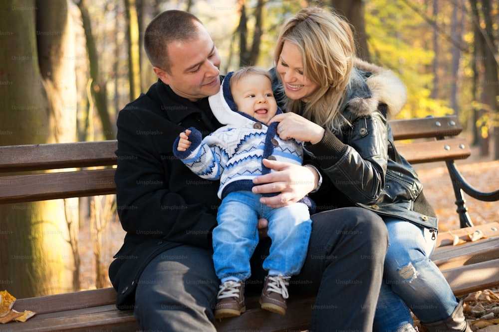 Familia joven, feliz y alegre, disfrutando al aire libre en un hermoso día de otoño. Follaje otoñal. Padre, madre y su hijito están sentados en un banco del parque y sonríen.