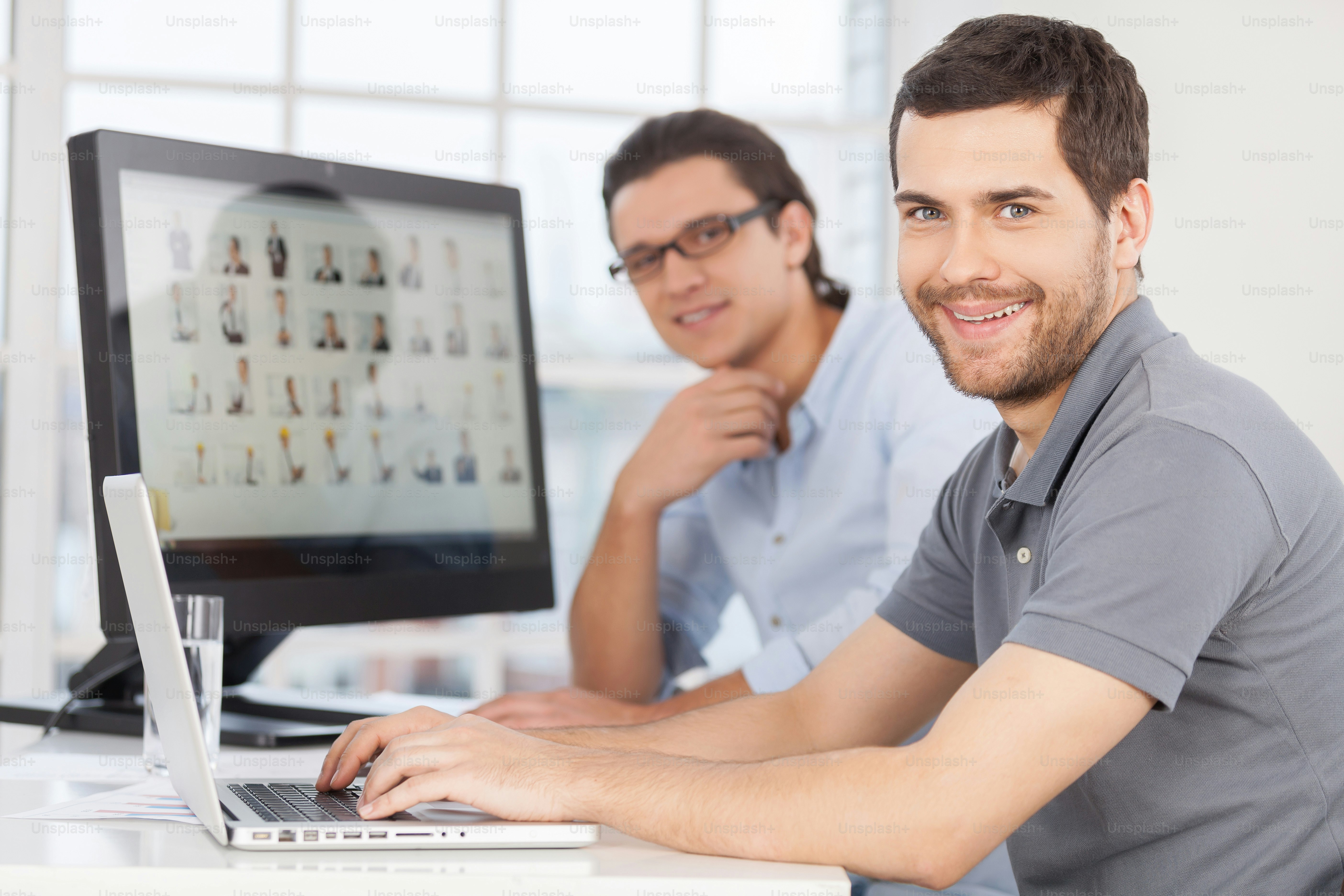 Two cheerful young men smiling at camera while sitting in front of computer monitors