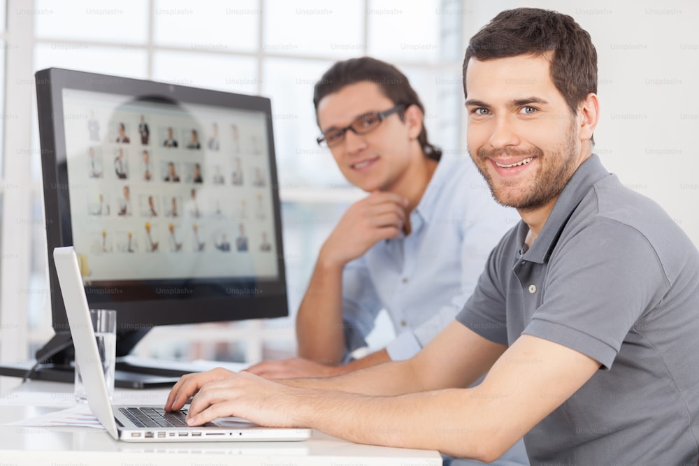 Two cheerful young men smiling at camera while sitting in front of computer monitors