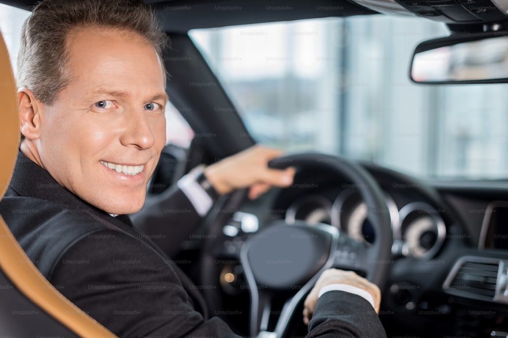 Rear view of confident senior man in formalwear sitting on drivers place in car and looking over shoulder