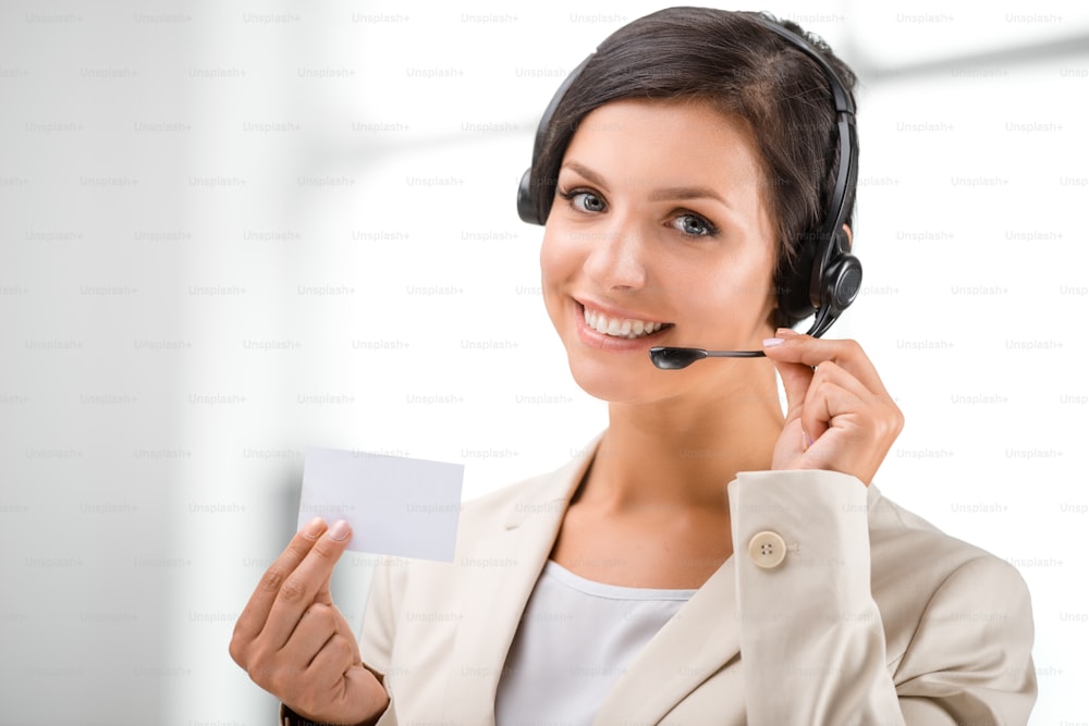 Beautiful smiling woman with headphones looking at camera and holding blank visit card at call center