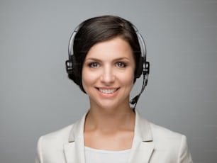 Beautiful smiling woman with headphones looking at camera at call center. Grey background