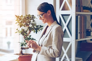 Side view of cheerful young beautiful businesswoman in glasses using her smart phone with smile while standing near window sill
