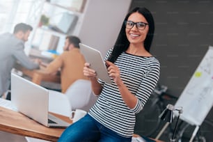 Jeune femme confiante tenant une tablette numérique et regardant la caméra avec un sourire tandis que ses collègues travaillent en arrière-plan