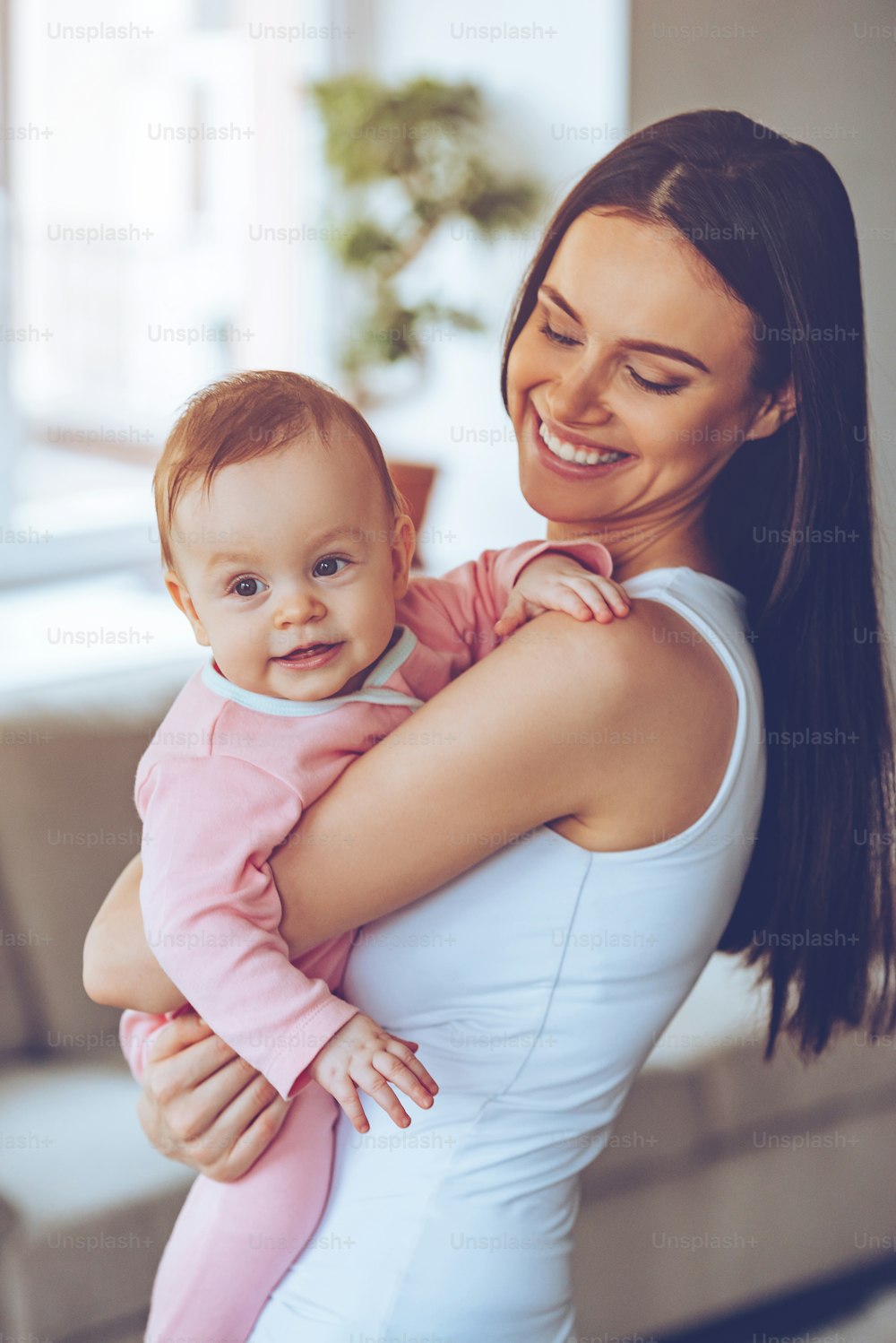 Beautiful young woman holding baby girl in her arms and looking at her with love while standing at home