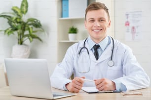 Ready to help. Cheerful pleasant positive pediatrician holding pen and expressing gladness while sitting at the table