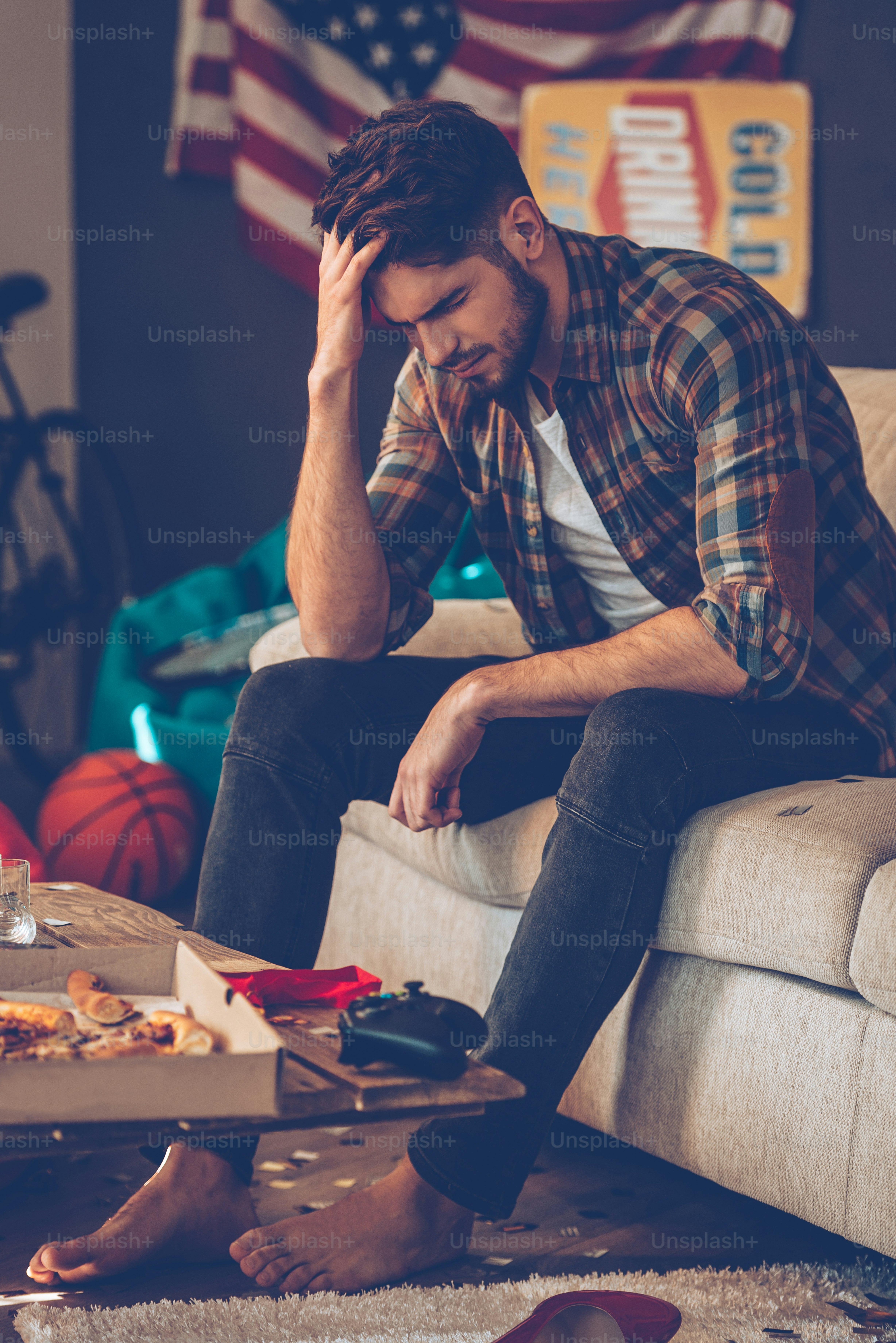 Frustrated young man keeping hand in hair while sitting on sofa in messy room after party