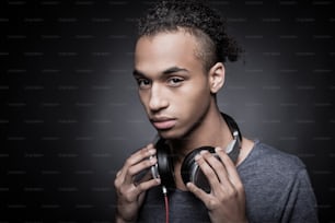 Portrait of young African man adjusting headphones and looking at camera while standing against black background