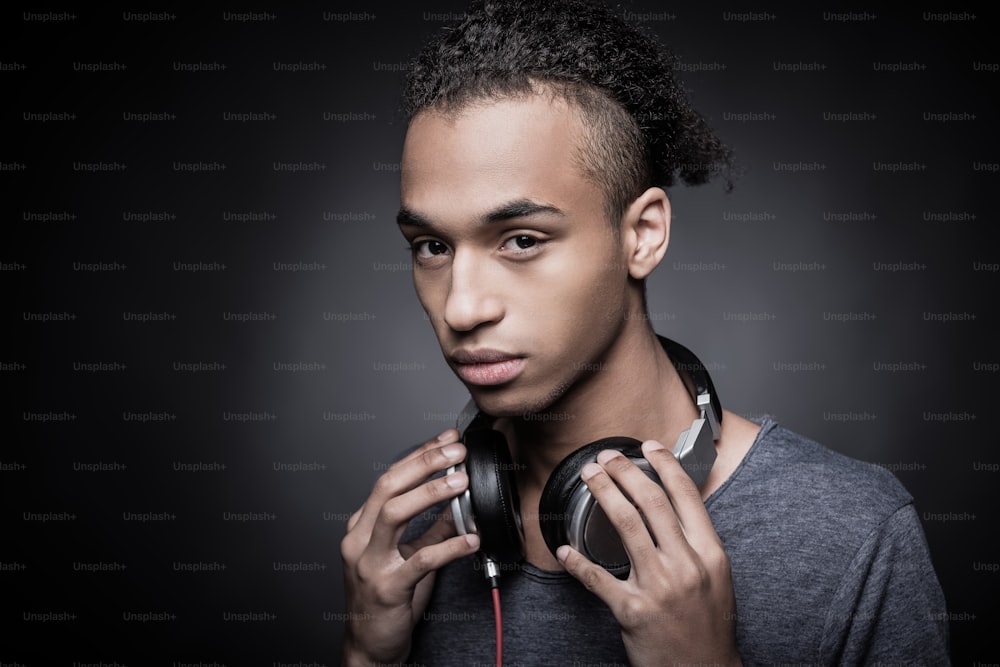 Portrait of young African man adjusting headphones and looking at camera while standing against black background