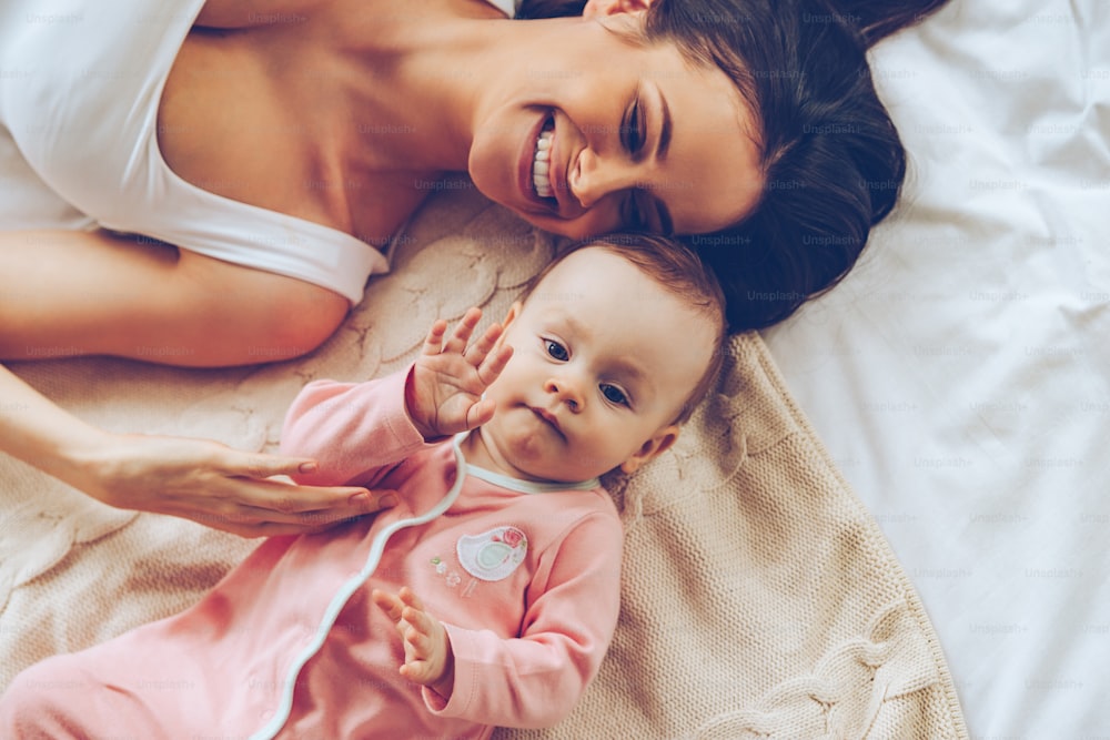 Top view of cheerful beautiful young woman playing with her baby girl while lying in bed
