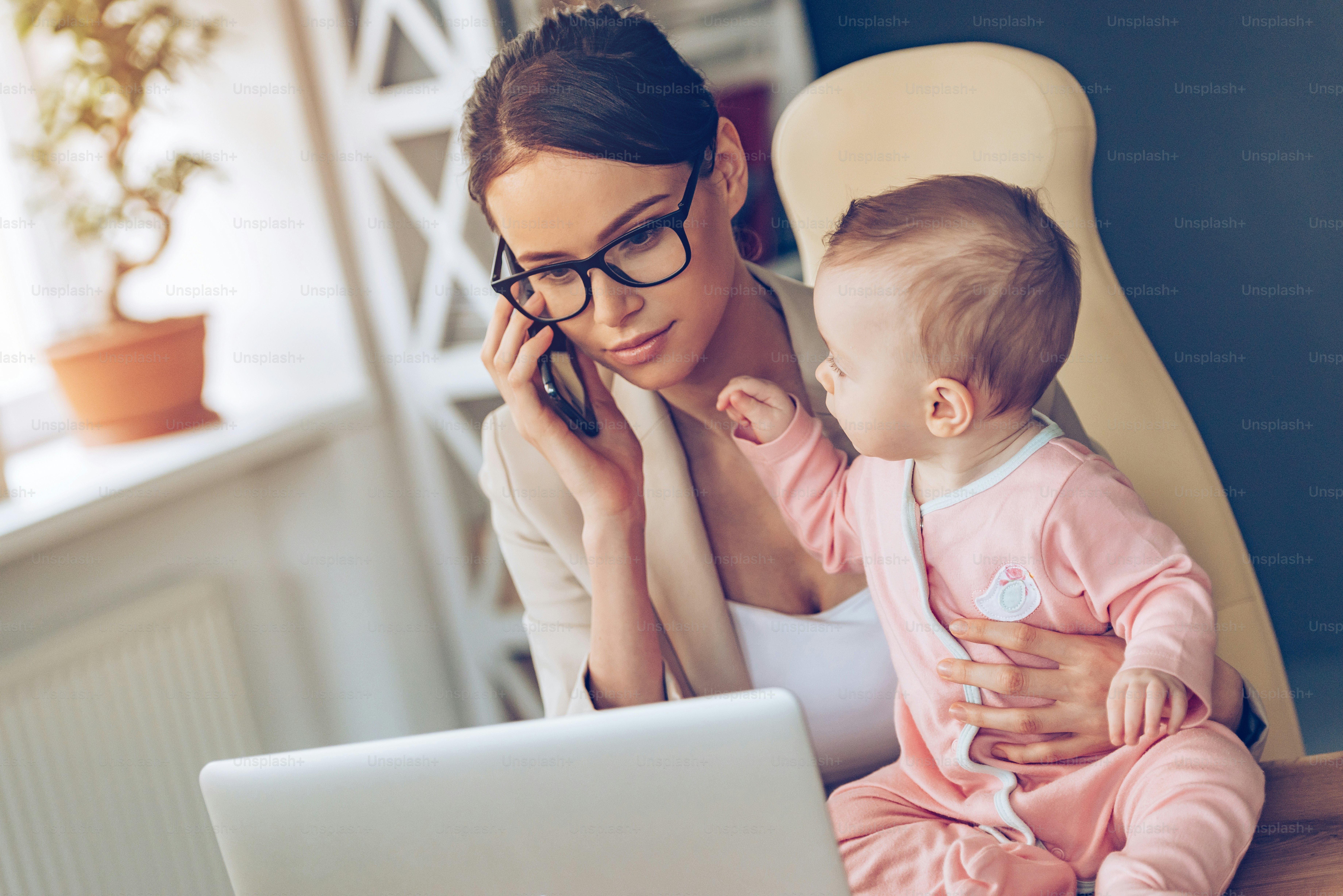 Young beautiful businesswoman talking on mobile phone and looking at laptop while sitting with her baby girl at her working place