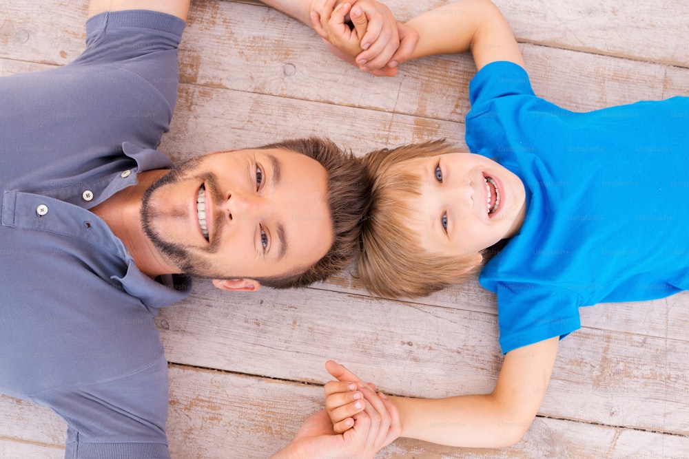 Top view of happy father and son holding hands smiling at camera while lying on the hardwood floor