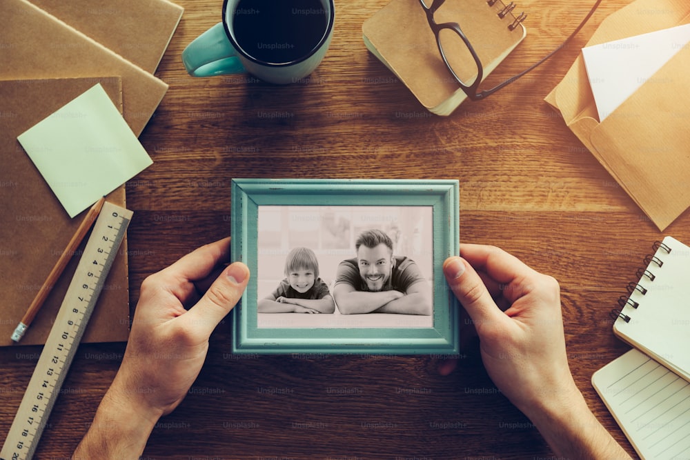 Close-up top view of man holding photograph of himself and his son over wooden desk with different chancellery stuff laying around