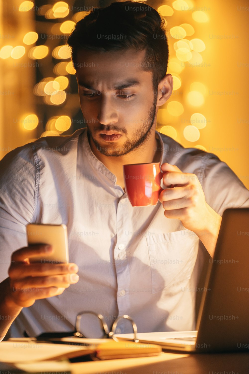 Joven seguro de sí mismo que mira su teléfono inteligente y sostiene una taza de café mientras está sentado en su lugar de trabajo por la noche con luces navideñas de fondo