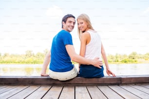 Beautiful young loving couple sitting at the quayside together and looking over shoulder with smile