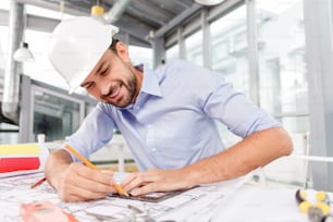 Un giovane ingegnere attraente sta disegnando schizzi di una nuova costruzione. Tiene in mano il righello e la matita. L'uomo è seduto al tavolo e sorride