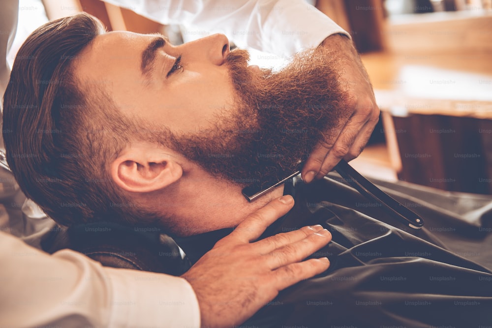 Vue latérale en gros plan d’un jeune homme barbu se faisant raser avec un rasoir à bord droit par un coiffeur