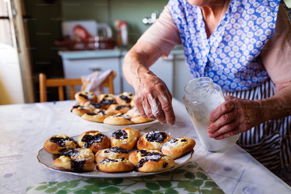 Donna anziana che cuoce torte nella sua cucina di casa. Cospargere i panini appena sfornati con zucchero a velo.