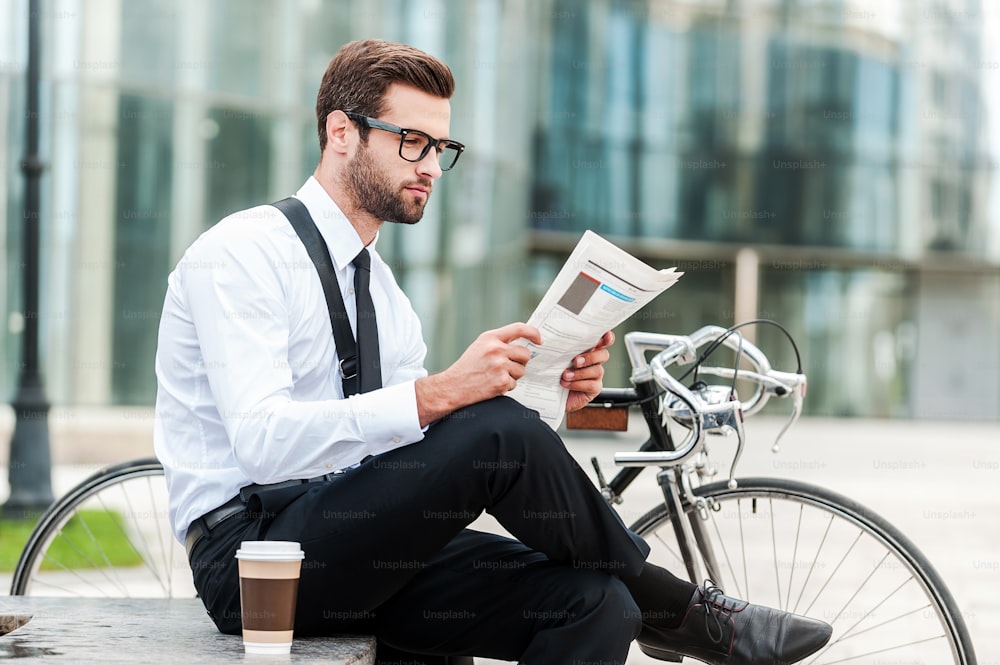Vista laterale di un giovane uomo d'affari che legge il giornale mentre è seduto vicino alla sua bicicletta con l'edificio per uffici sullo sfondo