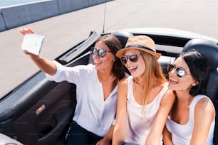 Vista superior de tres hermosas mujeres jóvenes que disfrutan de un viaje por carretera en convertible y hacen selfies