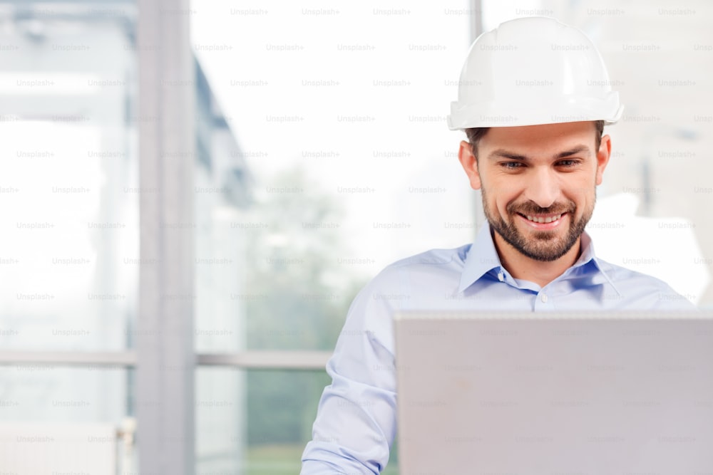 Handsome male engineer is using a notebook for work. He is sitting at the desk and smiling. The man is a white helmet is looking at the screen with aspiration. Copy space in left side