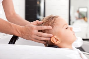 陽気な美容師の腕の接写。男性は髪を洗っている最中に男の子の頭にメッセージを送っています。子供は落ち着いていてリラックスしています