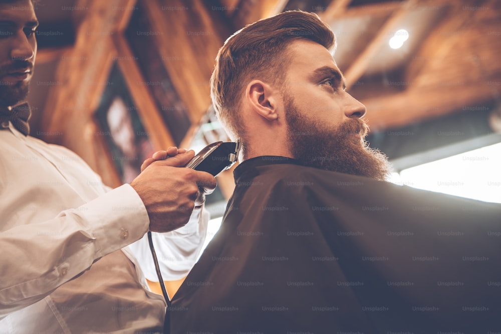 Vista ad angolo basso di un giovane barbuto che si fa tagliare i capelli dal parrucchiere con il rasoio elettrico dal barbiere