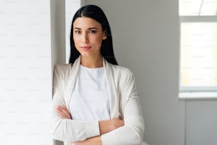 Beautiful young woman keeping arms crossed and looking at camera while standing near white wall