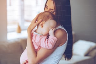 Side view of beautiful young woman holding sleepy baby girl in her arms and bonding with her while standing at home