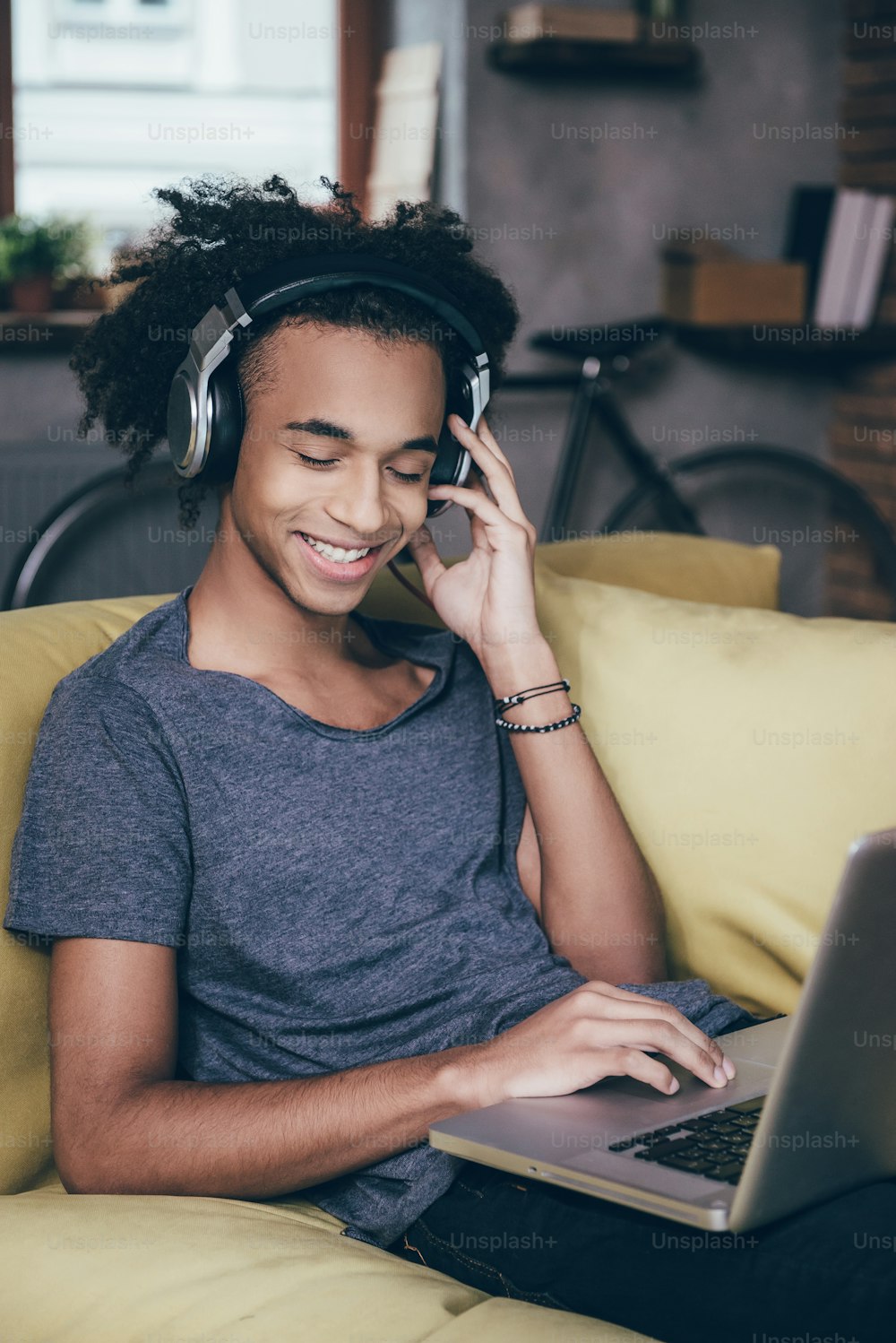 Cheerful young African man working on laptop and smiling while wearing headphones on the couch at home