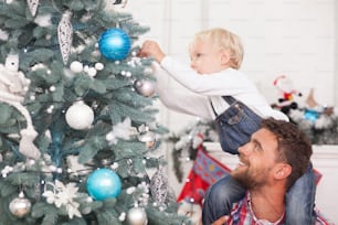 Padre e hijo alegres preparan el árbol de Navidad para la celebración. El chico está colgando la esfera con concentración. Su padre lo sostiene y mira feliz al niño. Está sonriendo