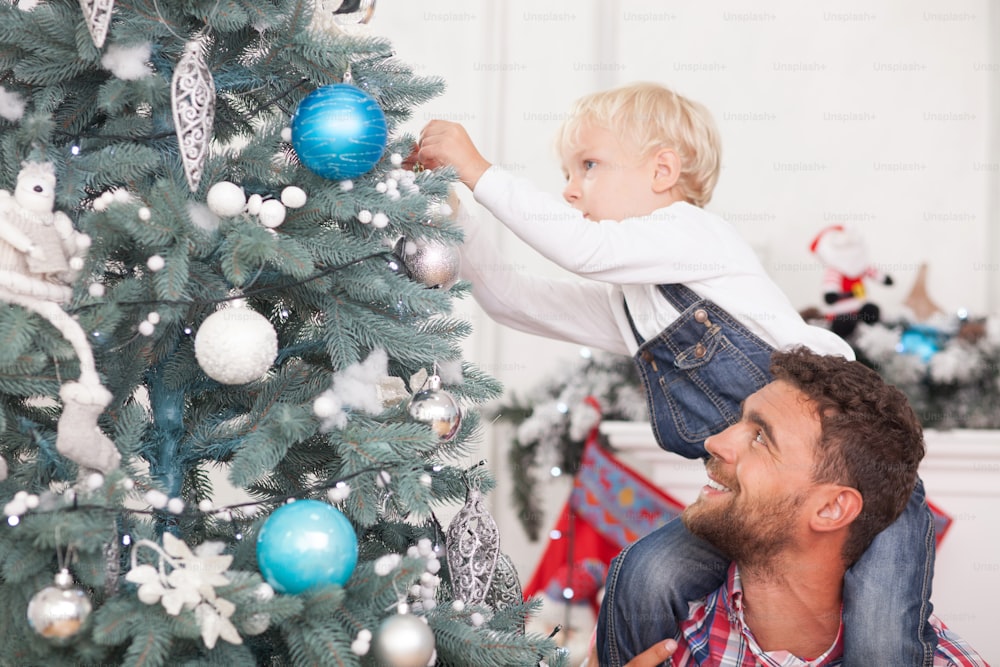 Fröhlicher Vater und Sohn bereiten den Weihnachtsbaum zum Feiern vor. Der Junge hängt Kugel mit Konzentration. Seine Eltern halten ihn fest und schauen das Kind glücklich an. Er lächelt