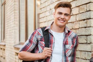Handsome young man carrying backpack on one shoulder and smiling while leaning at the brick wall