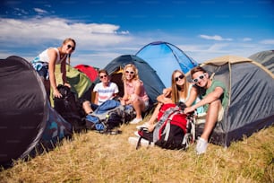 Group of teenage boys and girls at summer music festival, sitting on the ground in front of tents, packing